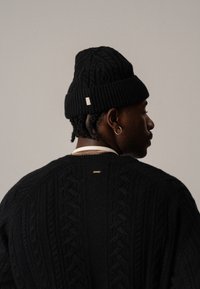 Zwarte gebreide beanie met een textuurpatroon, gedragen door een figuur in een bijpassende kabelgebreide trui met een kleine metalen accent aan de achterkant.