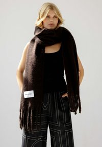 Écharpe en laine marron avec des franges et une étiquette rectangulaire indiquant "ARCTIC FOX." Portée lâchement autour du cou, avec une texture lisse.