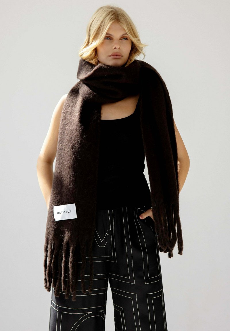 Écharpe en laine marron avec des franges et une étiquette rectangulaire indiquant "ARCTIC FOX." Portée lâchement autour du cou, avec une texture lisse.