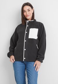 Black fleece jacket with a white chest pocket, white piping along the front, and side zip pockets. Features a high collar and elastic cuffs.
