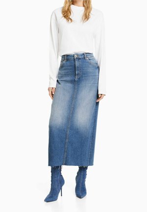 Jupe maxi en denim bleu clair avec un lavage décoloré, taille haute et ourlet effiloché, associée à des bottes à talons bleues assorties et un haut à manches longues blanc.