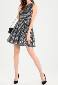 Zwart-wit gestreepte mouwloze jurk met een getailleerde bovenkant en een uitlopende rok, gecombineerd met een kleine zwarte handtas en hoge hakken.