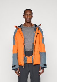 Άνδρας που φορά μια ανοιχτή πορτοκαλί και γκρίζα εξωτερική ζακέτα πάνω από ένα γκρίζο πουκάμισο με μακριά μανίκια και σκούρα γκρίζα παντελόνια, στέκεται μπροστά από έναν απλό φόντο.