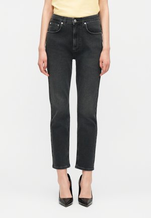 Femme portant un jean noir taille haute à jambes droites, associé à des escarpins noirs à bout pointu et un haut jaune pâle rentré.