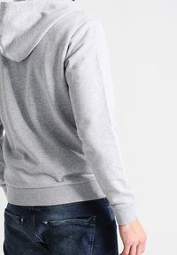 Felpa grigia con una texture morbida, dotata di cappuccio regolabile con stringhe e polsini a coste, abbinata a jeans scuri con dettagli sulle tasche.