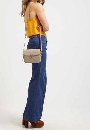 Bolso bandolera beige con cierre de solapa y herrajes plateados, llevado por una persona con camiseta amarilla y pantalones vaqueros acampanados azules, sobre un fondo blanco.