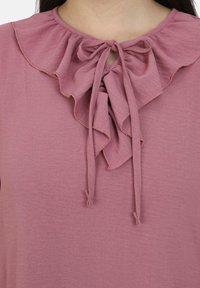 Blusa rosa con colletto arricciato, dotata di un dettaglio centrale con fiocco. Tessuto morbido e testurizzato con una leggera lucentezza.