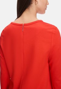 Rotes Langarmoberteil mit rundem Ausschnitt und einem dezenten Reißverschluss im Rücken. Glattes Material mit einer leicht taillierten Silhouette.