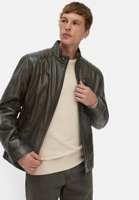 Herenjas van donkerbruin leer met een hoge kraag, gestikte details op de schouders en een ritssluiting aan de voorkant, gedragen over een lichtbeige trui.