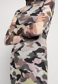 Femme portant une robe à manches longues et col montant avec un motif camouflage rose, beige, noir et vert olive, bras croisés sur la poitrine.