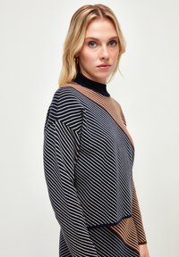 Sweter w paski w kolorach granatowym, brązowym i kremowym. Posiada wysoki kołnierz, ukośny wzór z bloków kolorów oraz teksturowany materiał. Standardowe dopasowanie.