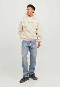 Kremfarget fleece hettegenser med snøring, med teksten "Originals Audio." Brukt over en lilla skjorte, sammen med lyseblå jeans og mørke sko.