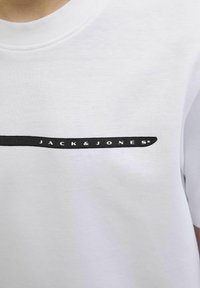 Balts kokvilnas T-krekls ar melnu horizontālu svītru, uz kuras rakstīts "JACK & JONES" baltām burtiem, ar apaļu kakla izgriezumu un īsām piedurknēm.