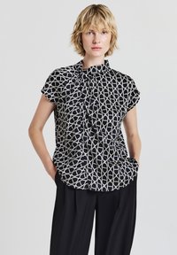 Blusa nera a maniche corte con un motivo geometrico bianco, caratterizzata da un colletto alto e un tessuto morbido, abbinata a pantaloni neri larghi.