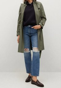 Femme portant un trench-coat vert olive, un pull noir, un jean bleu déchiré et des mocassins en cuir marron foncé, debout avec une main dans la poche.