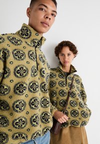 Pulovere din fleece de culoare verde măslin cu un model floral, guler înalt, închidere cu fermoar, croială lejeră, purtate de două persoane. Una dintre ele ține o geantă maro.