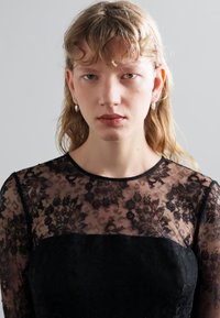 Schwarz gefärbtes Kleid mit durchsichtigen, floralen Spitzenärmeln, hohem Kragen und taillierter Passform. Glatte Textur mit aufwendigen Details im ganzen Kleid.