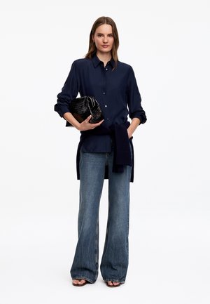Femme portant une chemise bleu marine à boutons et un jean à jambes larges, tenant une pochette noire texturée, avec un pull bleu marine noué autour de la taille.