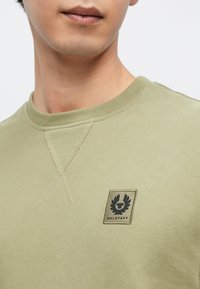 Olivengrønn sweatshirt med en ribbestrikket rund hals, synlig korsstingdetalj, og en rektangulær logo lapp med Belstaff-emblemet.