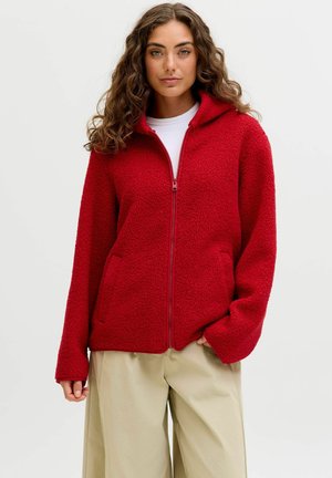 Frau mit langem lockigem Haar, die eine rote Fleecejacke mit Reißverschluss und Taschen sowie beigefarbene weite Hosen vor einem einfarbigen Hintergrund trägt.