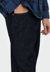 Jean en denim bleu foncé avec des détails de couture orange, présentant une poche sur le côté. Porté avec une chemise à carreaux bleue.