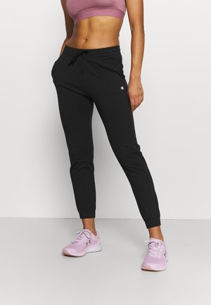 Mujer con pantalones jogger negros, sujetador deportivo rosa y zapatillas de correr rosas, de pie frente a un fondo blanco.
