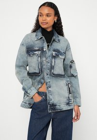 Svetlo modra, prevelika denim jakna z kontrastnimi temnejšimi madeži, štirimi sprednjimi žepi in žepom na rokavu z zadrgo. Črna tekstilna etiketa na strani.
