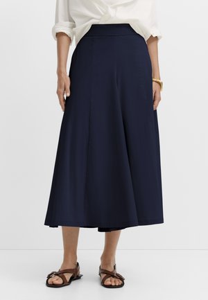 Frau trägt marineblaue, weit geschnittene, gekürzte Hose, weißes Hemd mit hochgekrempelten Ärmeln, braune Sandalen und goldenes Armband, steht vor weißem Hintergrund.