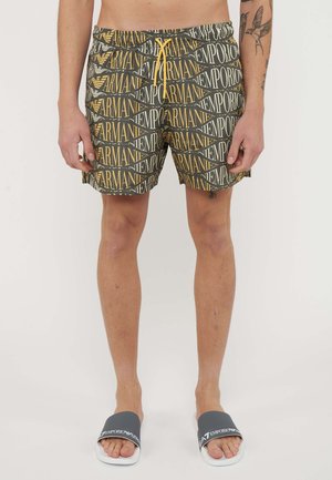 Modello maschile che indossa pantaloncini neri e oro Emporio Armani e sandali slides neri con suola bianca e logo Emporio Armani.