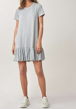 Femme portant une robe gris clair à manches courtes avec un ourlet à volants et des baskets blanches, debout devant un fond beige uni.