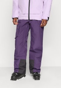 Pantalones de esquí impermeables morados con un bolsillo lateral, detalles grises en los puños y un corte ajustado, combinados con botas de esquí negras.