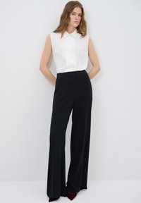 Witte mouwloze blouse met een kraag en gecombineerd met hoge zwarte wijde broek. Model draagt bordeauxrode puntige hakken.
