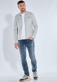 Hellgraue Zip-Jacke mit zwei Brusttaschen, über einem weißen Hemd getragen. Blaue Jeans und grau-weiße Sneakers runden das Outfit ab.
