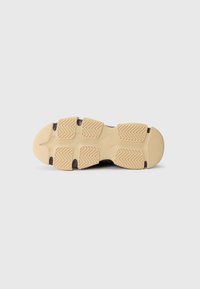 Suela de zapato deportivo con una base de goma beige texturizada, que presenta un patrón en zigzag para tracción y acentos oscuros a lo largo de los bordes.