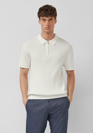 Jeune homme aux cheveux courts et bouclés portant un polo blanc à manches courtes côtelé et un pantalon bleu foncé, debout les mains dans les poches.