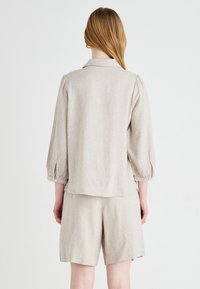 Completo in lino beige chiaro composto da una camicia con colletto e maniche a sbuffo, abbinato a pantaloni corti, che mette in mostra una texture naturale e un fit rilassato.