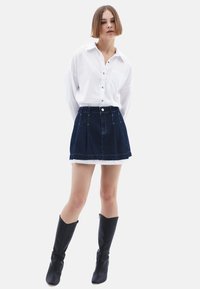 Donkere blauwe denimrok met plooien, rafelige zoom, gecombineerd met een witte blouse en kniehoge zwarte laarzen, op een witte achtergrond.