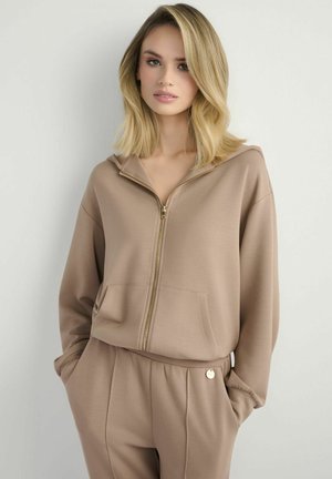 Blondynka w beżowej bluzie z zamkiem i dopasowanych spodniach, z rękami w kieszeniach, stoi na tle jasno szarego tła.