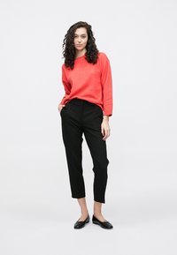 Koraļļu krāsas garroku džemperis ar brīvu piegriezumu, kombinēts ar melnām augsta jostasvietas biksēm ar šaurām kājām un melnām dzīvokļu kurpēm ar loku dekoru.