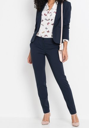 Femme portant un blazer bleu marine et un pantalon, une blouse blanche avec un imprimé papillon, des talons rose pâle et un sac bandoulière assorti.