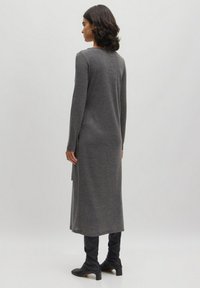 Lange, grijze trui-jurk van een zachte gebreide stof, met een rechte snit, lange mouwen en zijsplitten aan de zoom. Gemaakt in combinatie met zwarte laarzen.