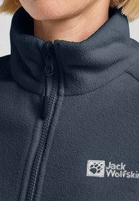 Giacca in pile grigio scuro con colletto alto, zip frontale e un logo con impronta di zampa. Texture morbida e senza motivi visibili.