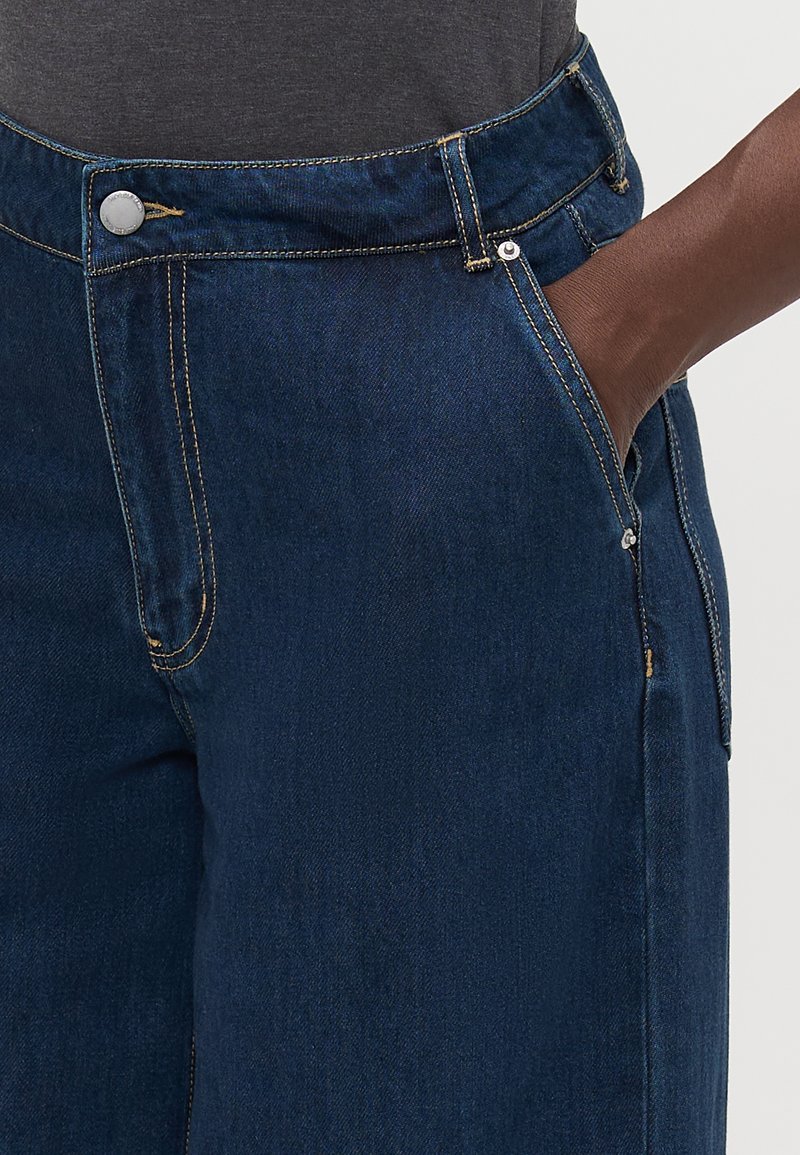 Ciemnoniebieskie dżinsy mają wysoki stan, kontrastowe przeszycia oraz zapięcie na guzik, a także kieszenie z przodu i gładką fakturę.