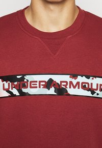 Primo piano di una persona che indossa una felpa rossa Under Armour con una striscia logo in camouflage bianco e nero che attraversa il petto.