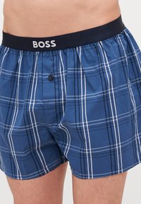 Modri karo bokserice z mornarsko elastično pasom z logotipom "BOSS", sprednjim gumbom in mehko bombažno teksturo. Črte v beli in temno modri barvi.