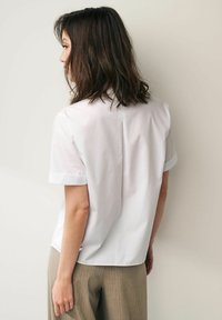 Chemise blanche à manches courtes avec une coupe droite, présentant un détail de couture au dos. Associée à un pantalon texturé marron clair.