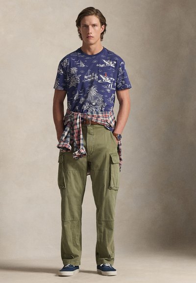 Μπλε-ναυτικό T-shirt με άσπρο τύπωμα, χακί παντελόνι cargo και καρό πουκάμισο δεμένο στη μέση. Μπλε αθλητικά παπούτσια ολοκληρώνουν το ντύσιμο.