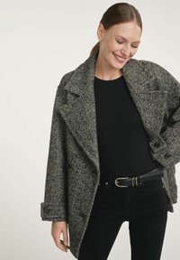 Kobieta uśmiechająca się, ubrana w czarną bluzkę i spodnie, z szarym wełnianym płaszczem w wzory zarzuconym na ramionach, na jednolitym tle.