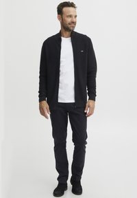 Fekete cipzáros kardigán bordázott textúrával, fehér póló felett viselve. Sötét nadrággal és sötét bokacsizmával párosítva. Egyszerű dizájn, minta nélkül.