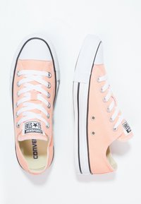 Des baskets en toile rose pastel avec semelles et bouts en caoutchouc blancs. Elles comportent des lacets blancs et des accents noirs sur la semelle. Design classique.